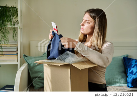 Serious upset young woman unpacking box with online purchases, looking at clothes 123210521