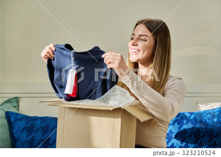 Young woman unpacking cardboard box parcel with online order, looking at new clothes 123210524
