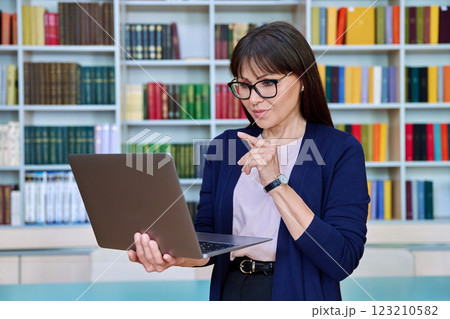 Portrait of middle aged serious woman teacher professor with laptop inside library Portrait of middle aged serious woman teacher professor with laptop inside library 123210582