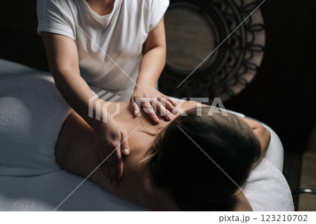 Close-up of professional female masseuse giving relaxing back massage to young unrecognizable woman lying on massage table in spa salon with dark interior, creating tranquil and soothing atmosphere. Close-up of professional female masseuse giving relaxing back massage to young unrecognizable woman lying on massage table in spa salon with dark interior, creating tranquil and soothing atmosphere. 123210702