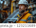 A bearded man in workwear and a hard hat is holding a cell phone 123211689