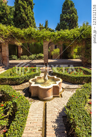 Alcazaba, palace and fortification in Malaga city at Andalusia, Spain, Europe. Serene courtyard garden with a stone fountain, meticulously manicured hedges, and brick pathways. A tranquil oasis. 123212158