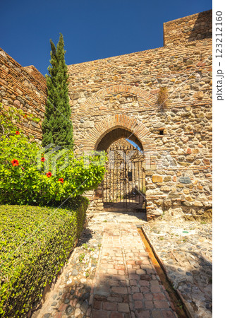 Alcazaba, palace and fortification in Malaga city at Andalusia, Spain, Europe. Ancient stone archway with iron gate, leading through a pathway bordered by lush greenery. Alcazaba, palace and fortification in Malaga city at Andalusia, Spain, Europe. Ancient stone archway with iron gate, leading through a pathway bordered by lush greenery. 123212160