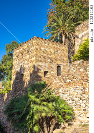 Alcazaba, palace and fortification in Malaga city at Andalusia, Spain, Europe. Ancient stone tower and wall, bathed in sunlight, with lush palm trees adding to the scene's serenity. 123212162