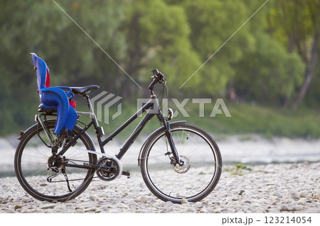 Close-up of new modern black bicycle with blue plastic child seat standing on side stand on lit by sun pebbles on blurred green trees summer bokeh background. Tourism and family traveling concept. 123214054