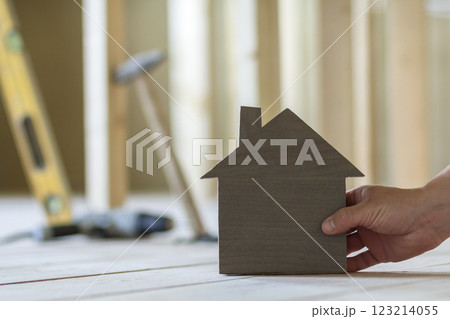 Close-up of muscular male hand holding small model house on blurred background of building tools in room under construction. Investments in real estate property and ownership of dream home concept. 123214055