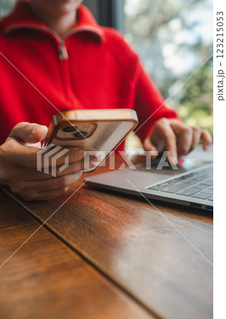 Close-up of a woman multitasking with a smartphone and laptop, wearing a red sweater, indoors. 123215053