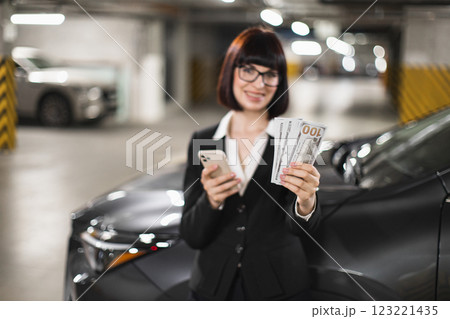 Caucasian business woman in formal attire holding cash and smartphone while standing in parking garage. Image represents success, technology, and finance in urban setting. Caucasian business woman in formal attire holding cash and smartphone while standing in parking garage. Image represents success, technology, and finance in urban setting. 123221435