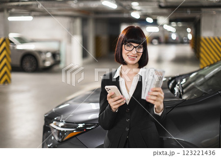 Caucasian businesswoman holding cash and smartphone in an internal parking lot, expressing confidence and success. Professional attire, positive expression. Caucasian businesswoman holding cash and smartphone in an internal parking lot, expressing confidence and success. Professional attire, positive expression. 123221436
