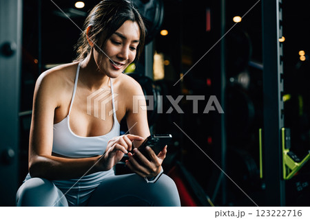 Asian woman resting on bench and checking all updates after exercising in gym interior, female using mobile phone application during training at fitness gym, sport workout healthy lifestyle concept 123222716