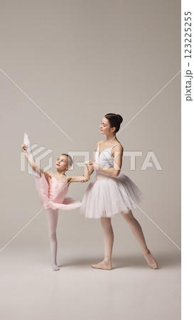 Little ballerina in pink tutu balances on one leg with arm extended, and mother helps her, showcasing concentration and grace against beige studio background. 123225255