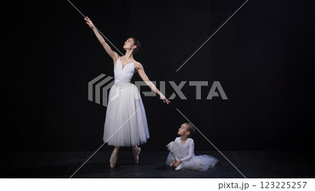 Adult ballerina pointing arm upward, child seated on floor looking up admiringly, both dressed in white ballet outfits, against black studio background. Adult ballerina pointing arm upward, child seated on floor looking up admiringly, both dressed in white ballet outfits, against black studio background. 123225257
