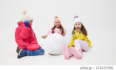 Three girls sitting near large snowball, laughing and preparing for next step in building snowman against white studio background. Three girls sitting near large snowball, laughing and preparing for next step in building snowman against white studio background. 123225266