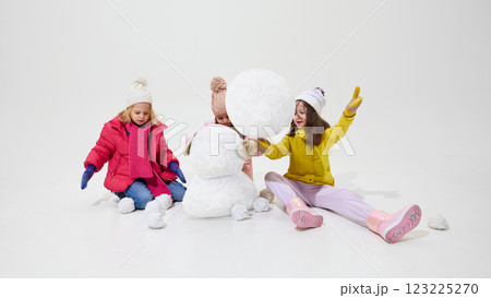 Three girls, having fun, placing final snowball on top, carefully shaping snowman while laughing against white studio background. 123225270