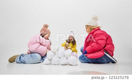 Three girls in colorful winter outfits sitting on floor, rolling snowballs and having fun together against white studio background. 123225278
