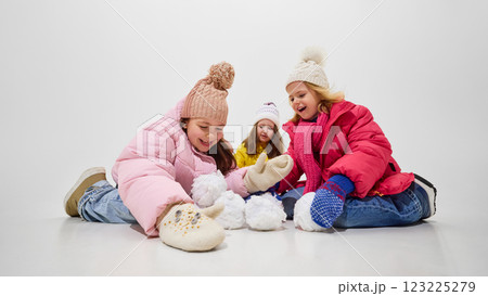 Girls in warm jackets playing with snowballs sitting on floor, laughing and having fun during winter game against white studio background. 123225279