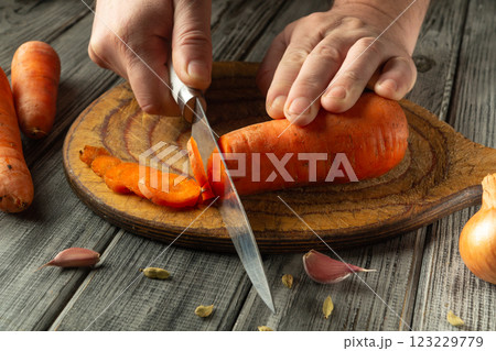 A person skillfully slices vibrant orange carrots on a wooden cutting board. Surrounding ingredients include garlic cloves and an onion, emphasizing preparation for a meal in a cozy kitchen A person skillfully slices vibrant orange carrots on a wooden cutting board. Surrounding ingredients include garlic cloves and an onion, emphasizing preparation for a meal in a cozy kitchen 123229779