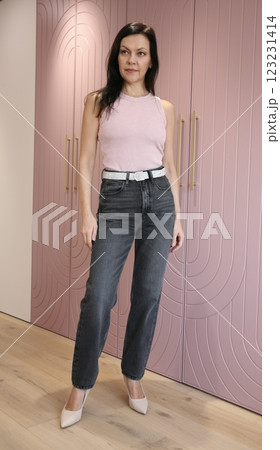 Pretty brunette 51 years old in pink high-heeled shoes in a pink fashionable T-shirt and grey jeans posing near a pink wardrobe 123231414
