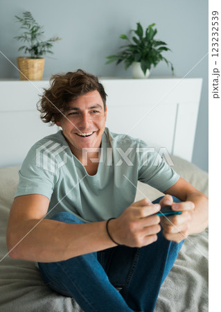 Handsome excited gamer sitting on a bed and playing video games on a console. Wireless controller and online game technologies concept 123232539