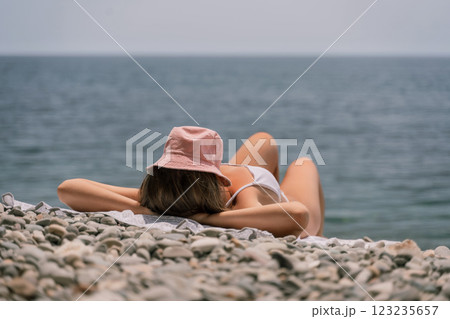 A woman is laying on the beach with a pink hat on her head. She is wearing a white bikini and is laying on a towel. The beach is rocky and the water is calm. Scene is relaxed and peaceful. 123235657