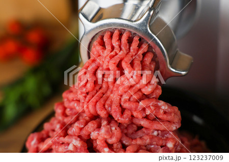 Concept of healthy and proper nutrition, cooking.Close-up of red ground beef with fat streaks coming out of a metal meat grinder on a wooden background, vegetables and herbs are visible out of focus 123237009