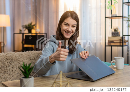 Smiling young woman freelancer closing laptop after work and showing thumbs up at table at home 123240175