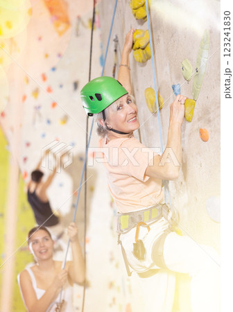 Positive senior old woman with rope training on concrete climbing rock wall to maintain healthy discipline in indoor adventure park Positive senior old woman with rope training on concrete climbing rock wall to maintain healthy discipline in indoor adventure park 123241830