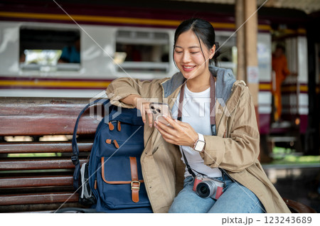 Smiling asian woman is looking through the phone while sitting on the wooden benchmark with bag. 123243689