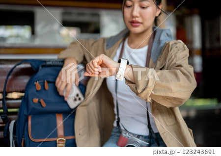 Asian woman looking at her wristwatch and holding the phone while sitting on the wooden benchmark. Asian woman looking at her wristwatch and holding the phone while sitting on the wooden benchmark. 123243691