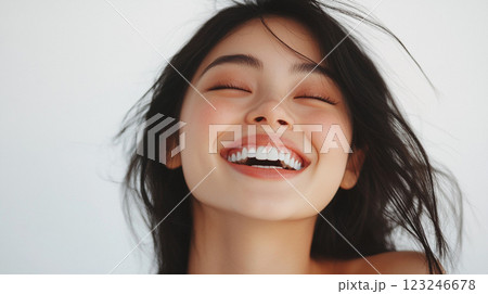 Joyful Asian woman with dark hair smiles brightly with closed eyes, radiating happiness and positive energy in a minimalist setting. 123246678