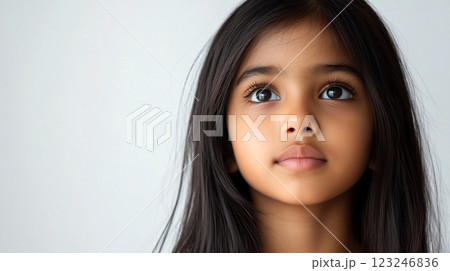 A young girl with long, straight dark hair looks upwards with a thoughtful expression against a simple, light-colored background. 123246836
