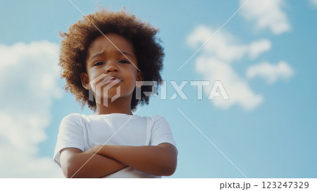 A young child poses with arms crossed against a bright blue sky. The confident look on their face gives an air of determination and self-assurance. A young child poses with arms crossed against a bright blue sky. The confident look on their face gives an air of determination and self-assurance. 123247329