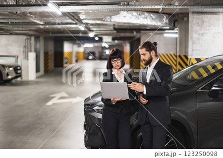Caucasian man and woman in business attire reviewing laptop documents in parking garage. Professional collaboration, teamwork atmosphere, urban setting. Both adults in suit, focused on task. Caucasian man and woman in business attire reviewing laptop documents in parking garage. Professional collaboration, teamwork atmosphere, urban setting. Both adults in suit, focused on task. 123250098