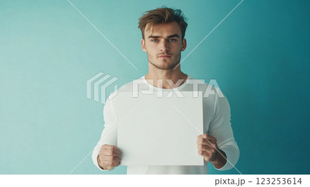 Young man holds a blank white sign in front of a blue background, space to add text or information. Studio shot of person with signage. 123253614