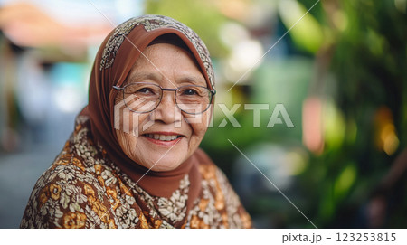 Portrait of a smiling older woman wearing a hijab and glasses, radiating warmth and kindness. She is set against a green leafy backdrop. 123253815