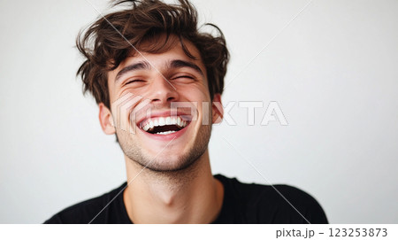 A joyful young man with curly brown hair and a black t-shirt is laughing with his eyes closed against a plain background. A joyful young man with curly brown hair and a black t-shirt is laughing with his eyes closed against a plain background. 123253873