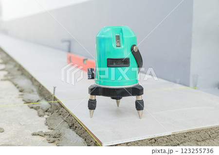 A green laser level on a tripod projects beams across a white and gray floor, ensuring precision while laying ceramic tiles in a construction setting. 123255736
