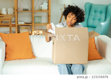 African girl unpacking delivery looking in box. Happy woman opening carton box. Female getting parcel looking at delivered goods items. Satisfied client positive feedback quick express deliver 123256976