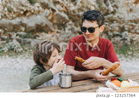 Close-up view of father hugs his school boy son on a family picnic. Child kid and his dad taking a rest and enjoying a picnic while hiking. Boy smiles and bites the bread donut on a picnic Close-up view of father hugs his school boy son on a family picnic. Child kid and his dad taking a rest and enjoying a picnic while hiking. Boy smiles and bites the bread donut on a picnic 123257364