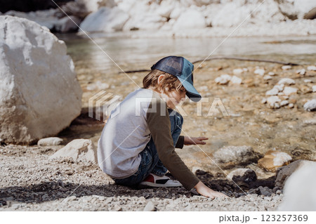 Caucasian school boy sits on a riverside rock in the canyon with mountain cliffs in the background. Kid child taking a rest on a boulder near mountain river bank Caucasian school boy sits on a riverside rock in the canyon with mountain cliffs in the background. Kid child taking a rest on a boulder near mountain river bank 123257369