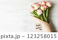 A bouquet of pink tulips wrapped in a light craft paper sits on a white wooden surface. A tag attached reads 'Happy Mother's Day,' celebrating the occasion with elegance. 123258015