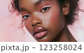A close-up portrait of a young Black woman with beautiful skin and stylish makeup. The makeup features pastel colors, bold eyeliner, and glossy lips. The background is soft pink, enhancing her 123258024