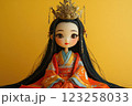 A beautiful traditional doll wearing a colorful robe and an ornate crown. The doll features long, black hair and a calm expression. The background is a solid yellow color. 123258033
