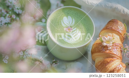 A vibrant green matcha latte rests in a cup adorned with floral latte art. Next to it lies a fresh, flaky croissant. The scene features soft pastel colors and natural light. 123258051