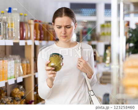 Girl customer in supermarket examines jar of canned pea, chooses food-stuff in grocery Girl customer in supermarket examines jar of canned pea, chooses food-stuff in grocery 123258460