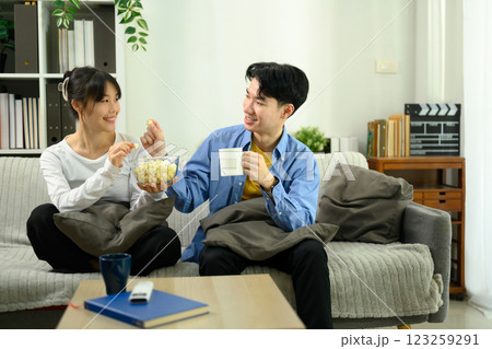 Happy young couple sitting on the couch, sharing a bowl of popcorn while watching movie Happy young couple sitting on the couch, sharing a bowl of popcorn while watching movie 123259291