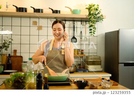 Smiling middle aged woman in apron cooking and talking on mobile phone in a kitchen Smiling middle aged woman in apron cooking and talking on mobile phone in a kitchen 123259355