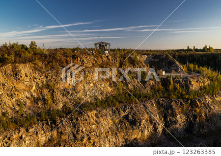A rocky cliff with sparse trees and dry bushes under warm sunlight. At the top, an abandoned industrial structure stands among autumn foliage, creating a contrast between nature and human-made ruins. 123263385