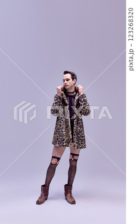 Artistic young man standing in leopard coat and fishnet tights with fierce expression and confident stance against white studio background. 123268320