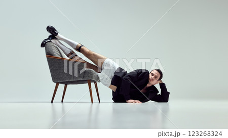 Expressive young man, dressed stylish outfit, leaning off chair with relaxed yet unusual posture against white studio background. 123268324